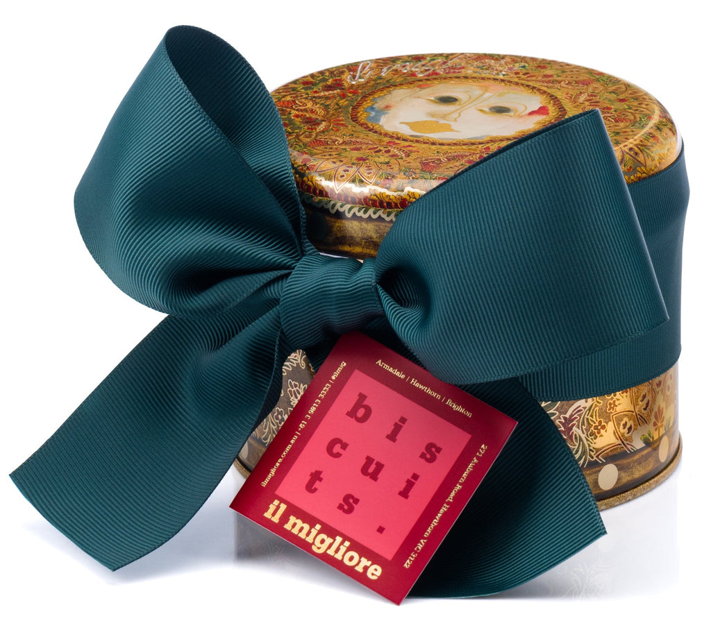 A round tin of biscotti wrapped in a large dark teal ribbon bow, with a red paper tag reading 'biscotti il migliore' attached. The tin has an ornate gold and red illustrated lid.