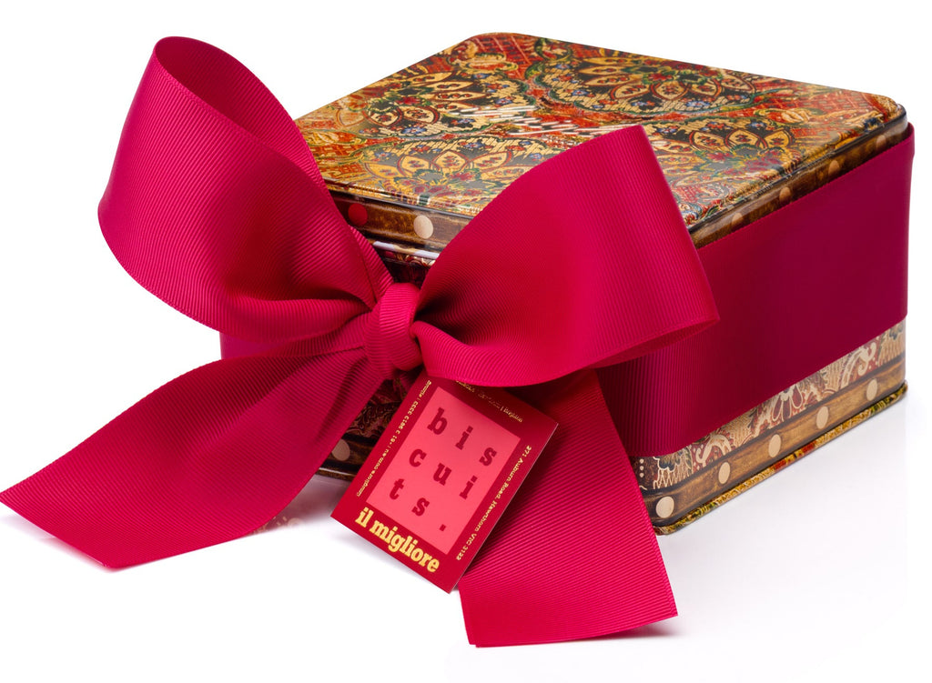A square gift box with a colorful ornate pattern lid and a wide pale red ribbon tied in a large bow, with a small red tag reading 'il migliore' attached, on a white background. Packed with delicious biscuits 