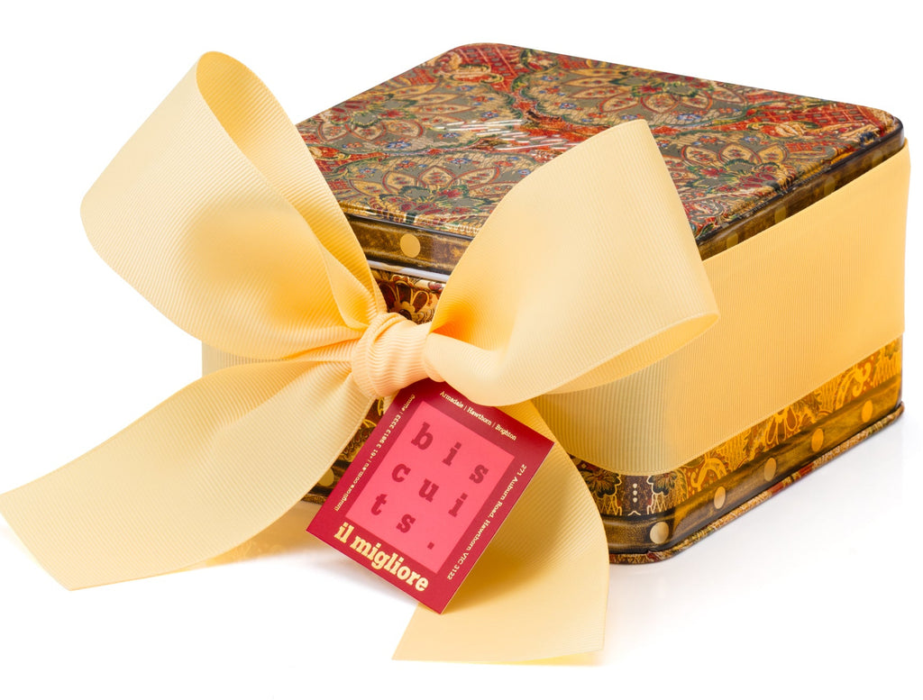 A square gift box with a colorful ornate pattern lid and a wide pale yellow ribbon tied in a large bow, with a small red tag reading 'il migliore' attached, on a white background.